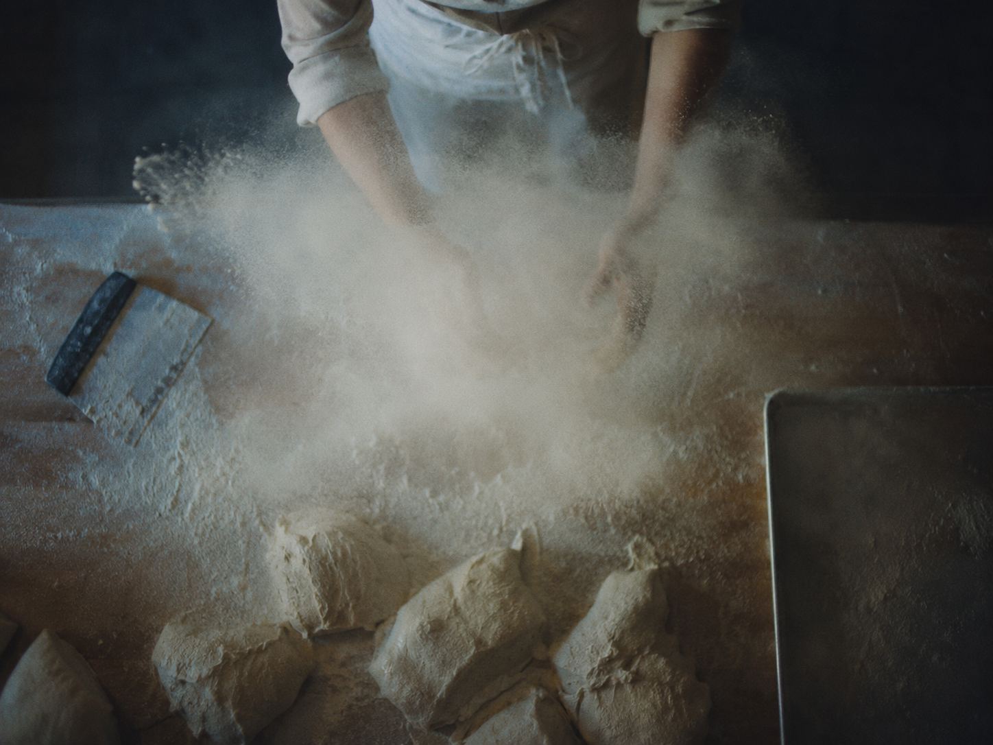 A baker throwing flour across a bench of shaped dough