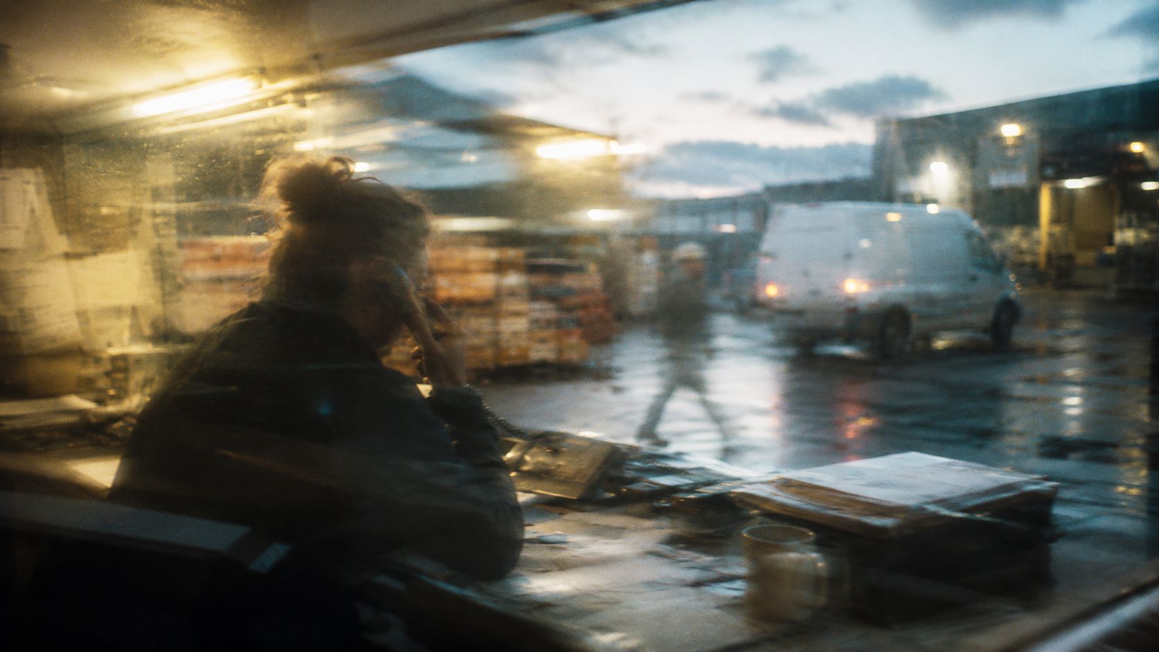 An order manager on the phone behind rain-streaked glass, the yard moving outside